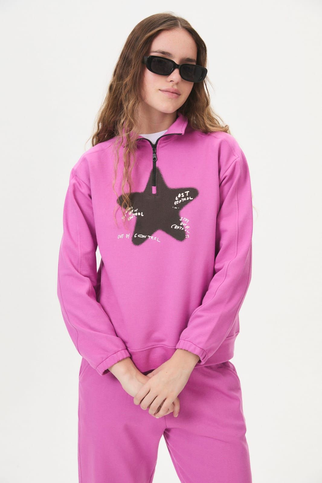 Mujer con sudadera rosa y pantalones con un gráfico de estrella negra, usando gafas de sol sobre un fondo blanco.