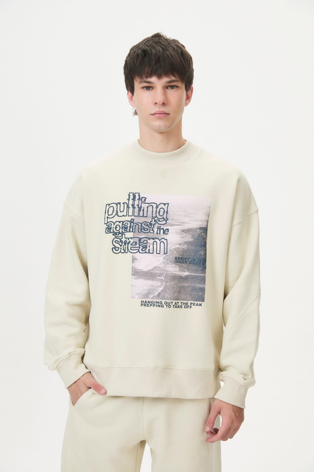 Modelo con sudadera color crema y pantalones a juego, de pie frente a un fondo blanco de estudio.