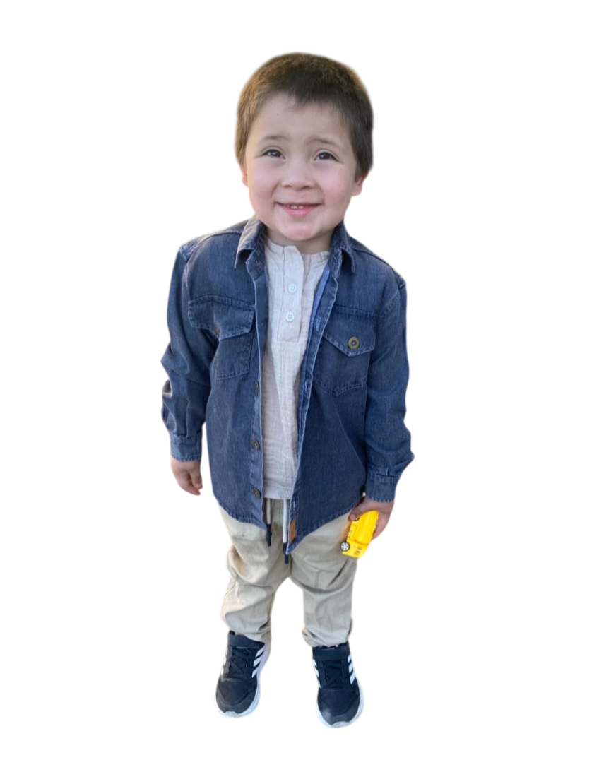 Niño sonriente con chaqueta vaquera y pantalones claros, de pie sobre un fondo blanco y sosteniendo un juguete amarillo.