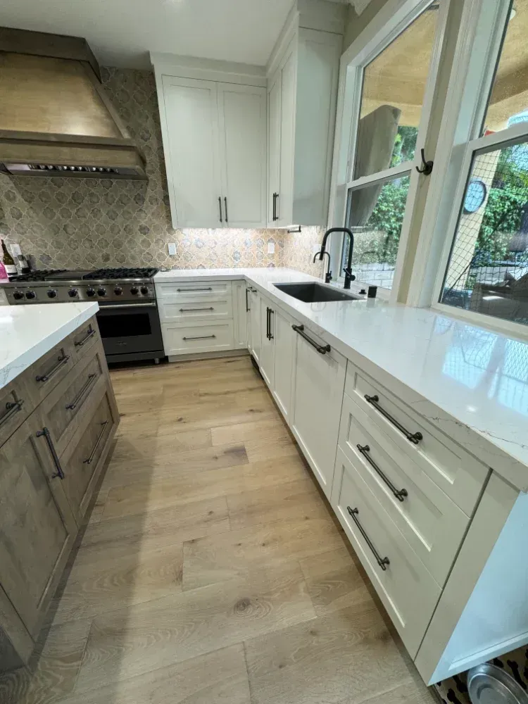 A bright kitchen with white cabinets, light wood flooring, and a black faucet. The room has natural light from a large window.