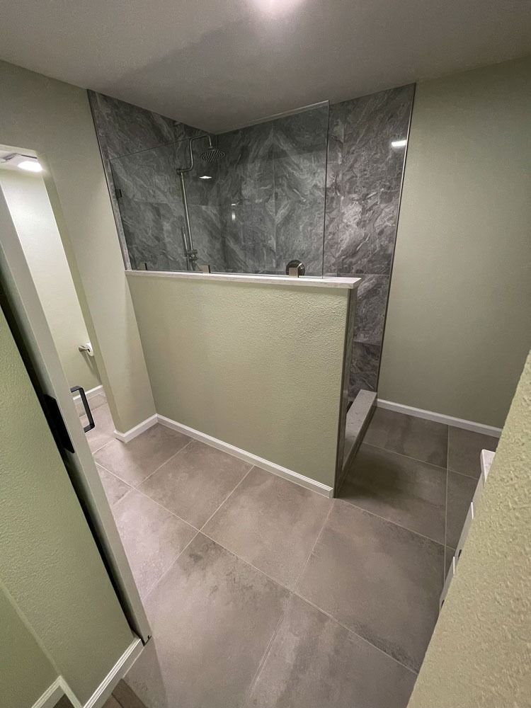 Bathroom with gray tile flooring and a walk-in shower featuring gray marbled tile. Light green walls and a white shower ledge.