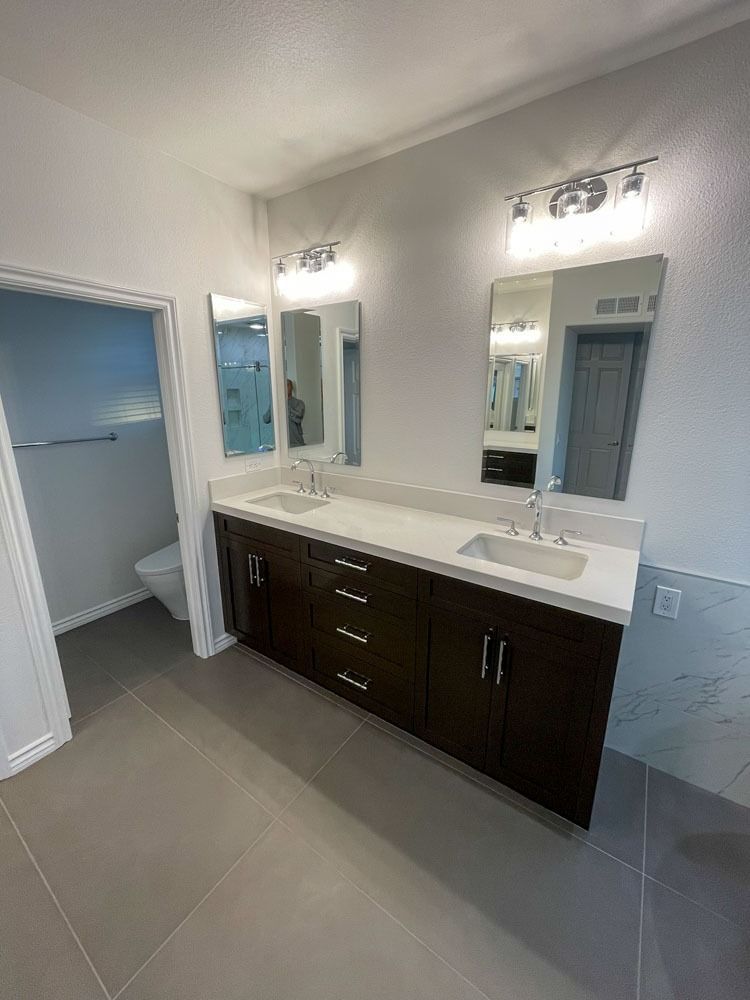 Bathroom with a dark wood double vanity, two rectangular mirrors, and gray tiled floors. The room includes a toilet and shower area.