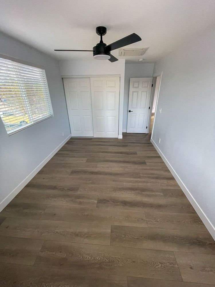 Empty bedroom with wood-look flooring, white trim, and pale blue walls.  White closet doors and a closed white door are visible. Black ceiling fan.