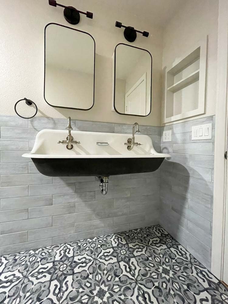 Bathroom with a black and white patterned floor, gray tiled walls, a white sink, two mirrors, and a built-in shelf.