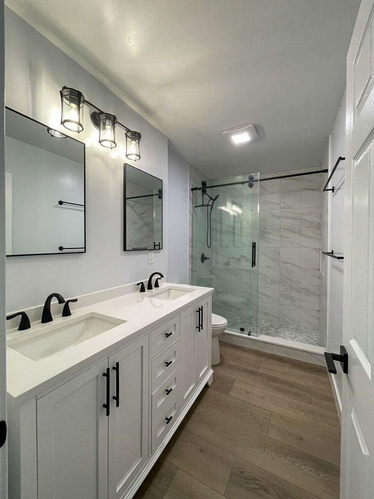 A renovated bathroom featuring a white vanity with two sinks, a glass-enclosed shower with marble-like tiles, and light wood-look flooring.