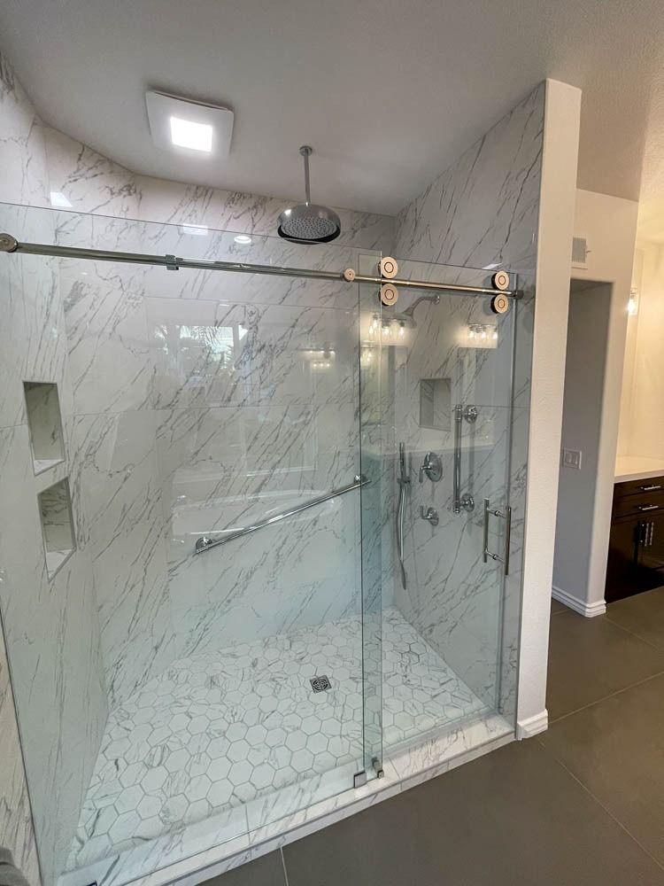 A modern, spacious shower with marble-like tiled walls and a sliding glass door. Gray tile flooring and silver fixtures complete the elegant bathroom.