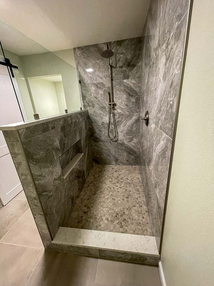 A gray tiled walk-in shower with a pebble stone floor. It has a built-in shelf, a rain shower head, and is partially enclosed by a wall.