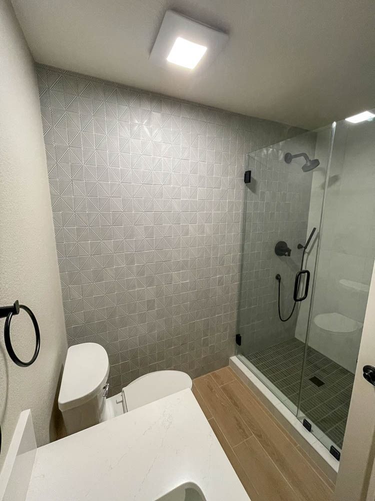 A modern bathroom with gray tile walls, a glass shower, a white toilet, and a wooden floor.
