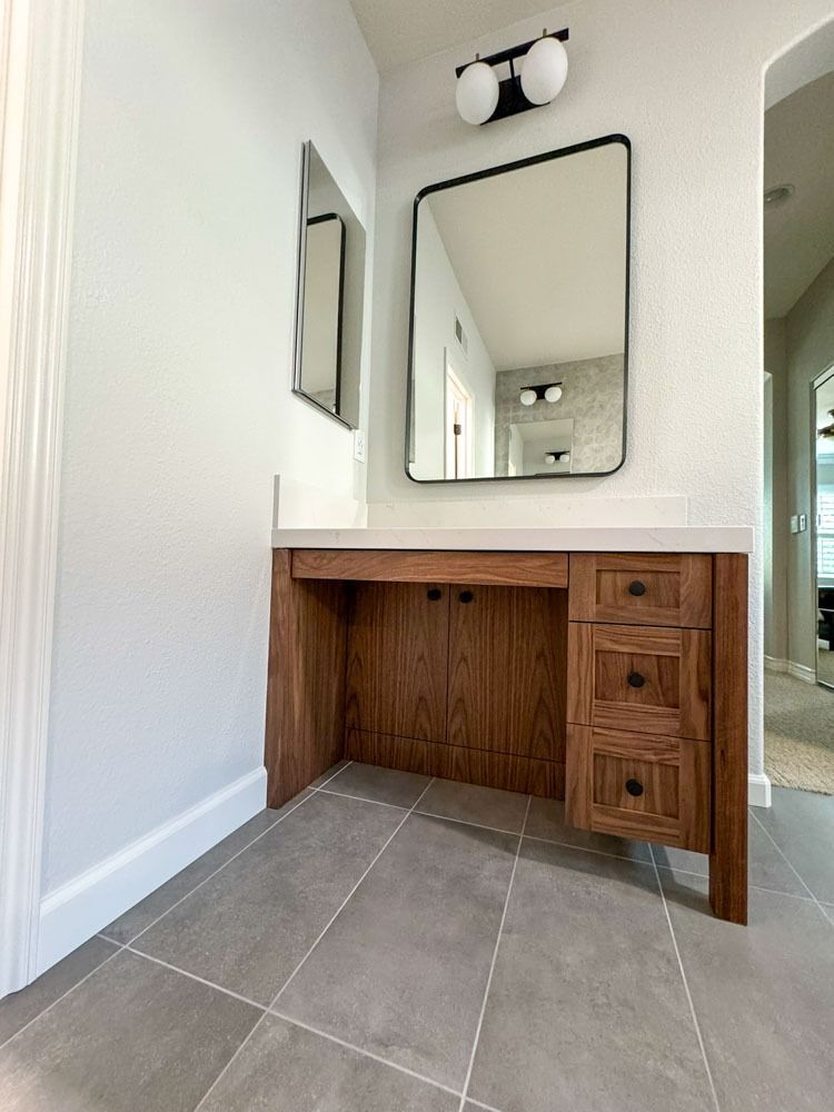 A wooden vanity with a mirror and light fixture in a bathroom. The vanity has a cabinet and drawers. Gray floor tiles.