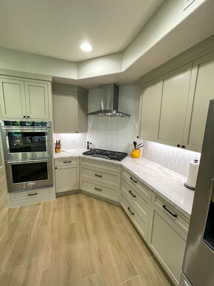 A modern kitchen with light-colored cabinets, countertops, and wood-look flooring. Stainless steel appliances and a range hood are featured.