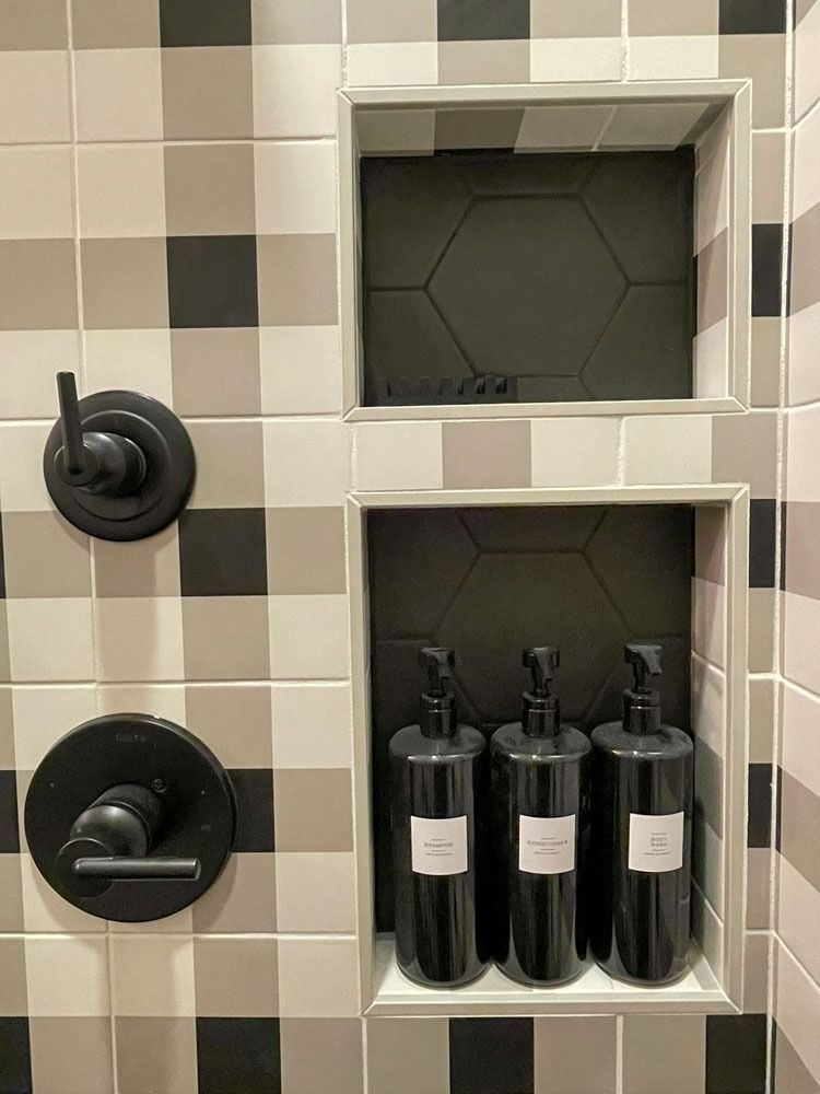 Shower with black fixtures and built-in shelves holding black pump bottles, set in a checkered gray and white tile wall.