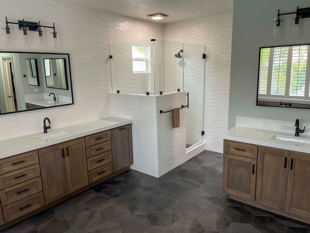 A modern bathroom with two vanities, a glass shower, white textured walls, and dark gray hexagonal tile flooring.