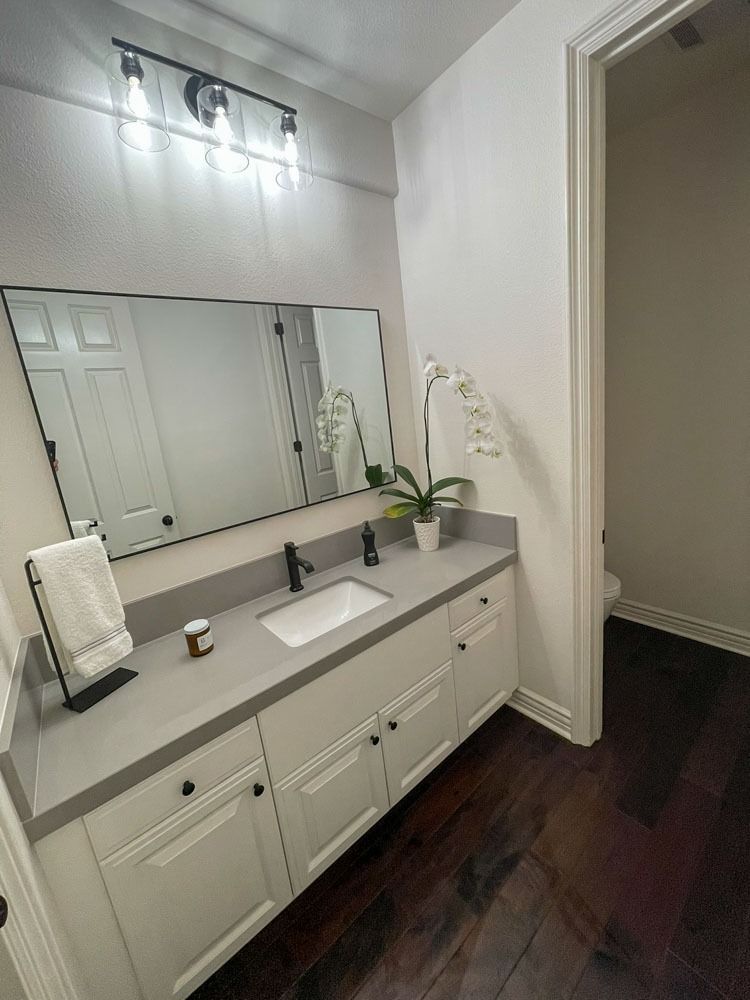 Bathroom with white cabinets, gray countertop, and a large mirror. Dark wood floor and a door to a toilet.