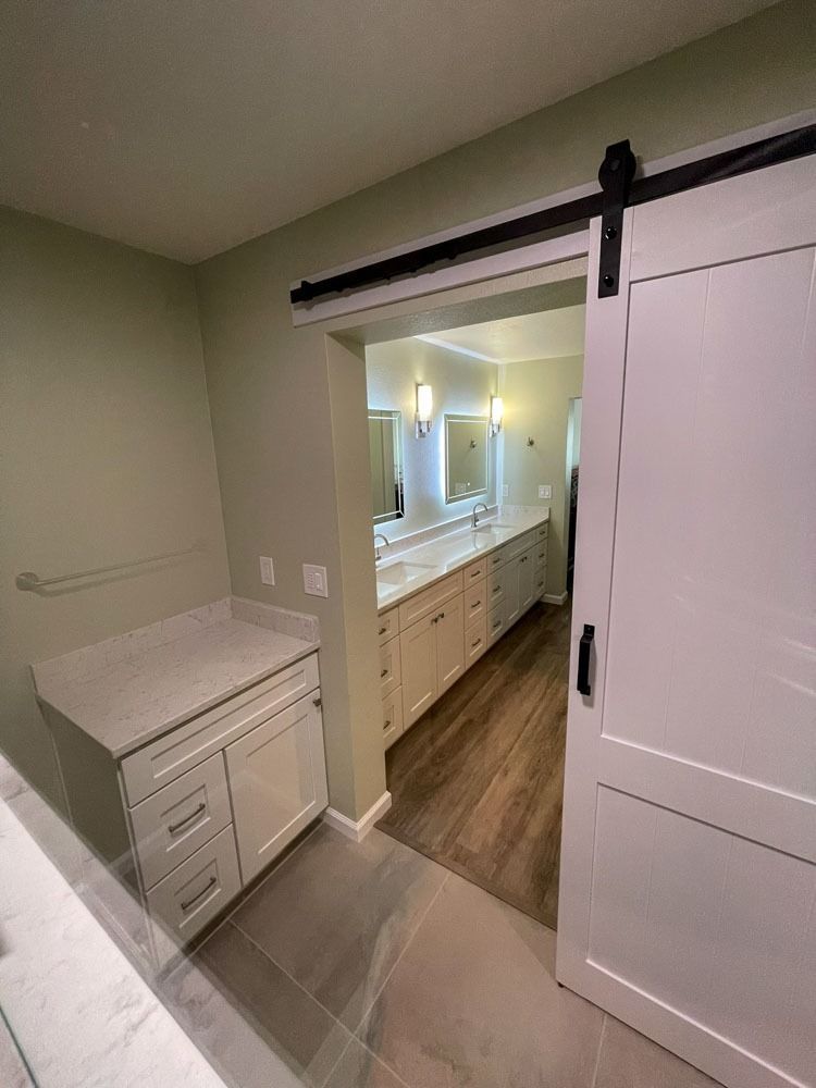 A white barn door partially open revealing a bathroom with a long white vanity and light green walls. A small white cabinet is on the left.