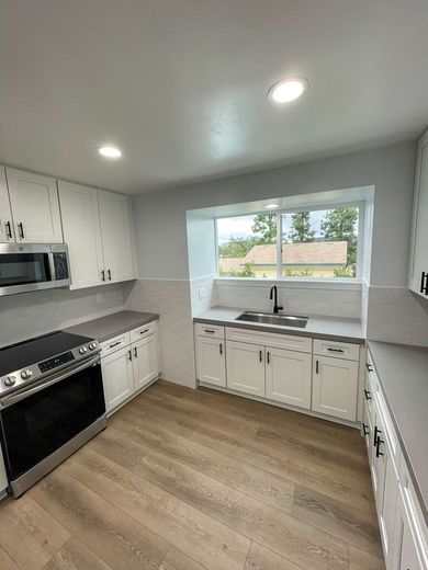 Modern white kitchen with stainless steel appliances, gray countertops, and light wood flooring; a window offers an exterior view.
