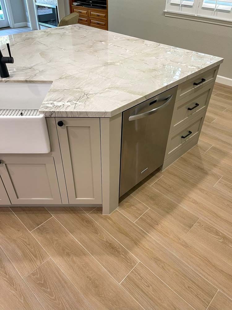 Kitchen island with light-colored countertop, light gray cabinets, stainless steel dishwasher, and white farmhouse sink, situated on light wood-look flooring.