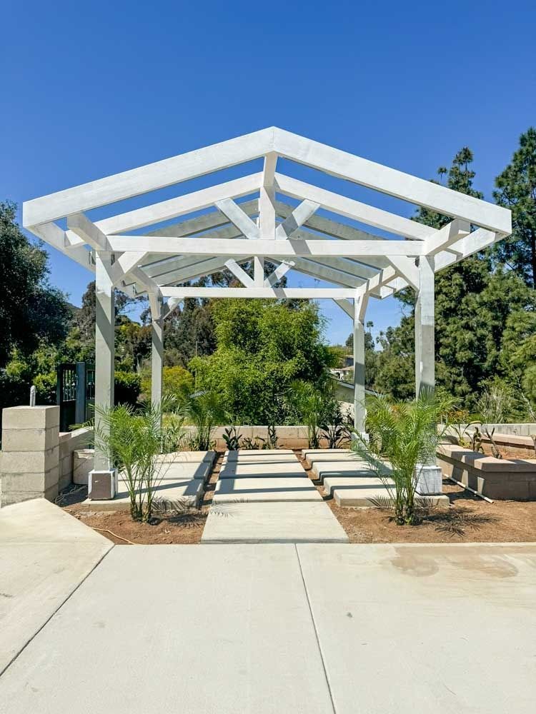 White pergola in a garden setting with a pathway leading to it. Bright blue sky and green trees are visible.