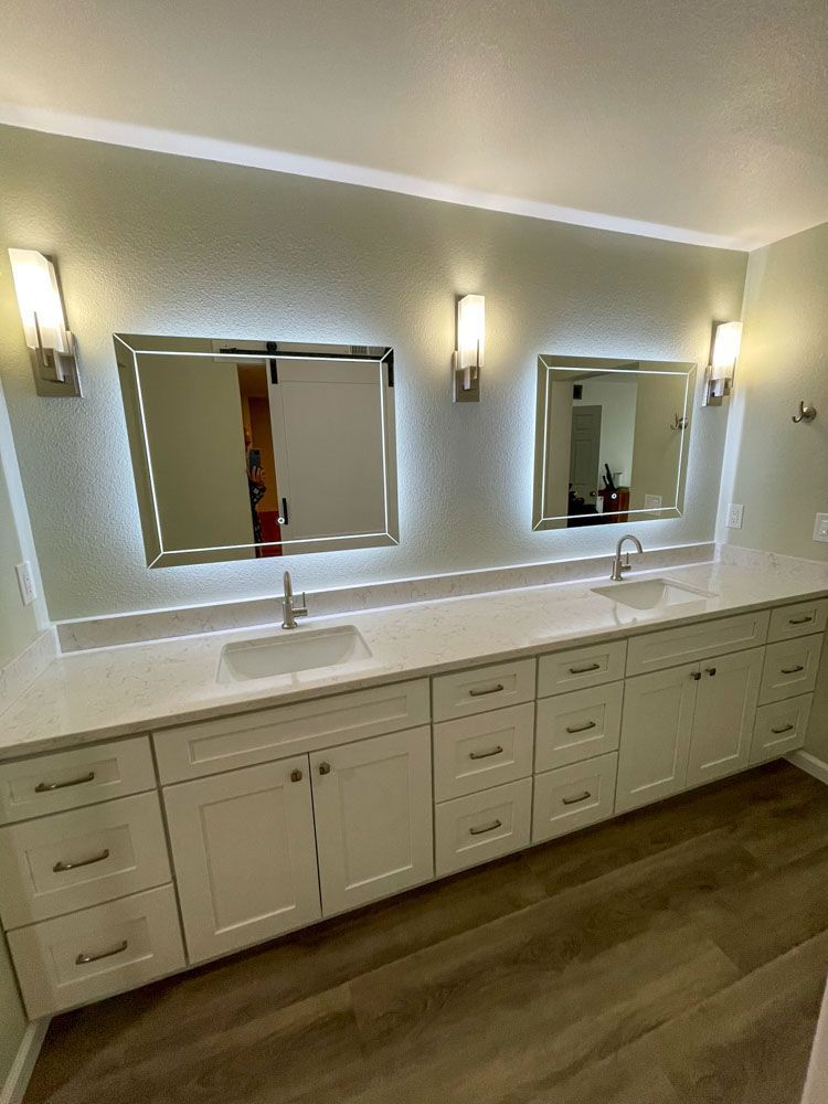 A modern bathroom vanity with two illuminated mirrors, white cabinets, and silver fixtures against a light green wall.