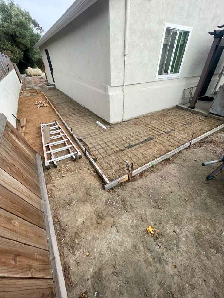Concrete patio under construction next to a building. Rebar framework sits inside wooden forms on a dirt surface, with a ladder nearby.
