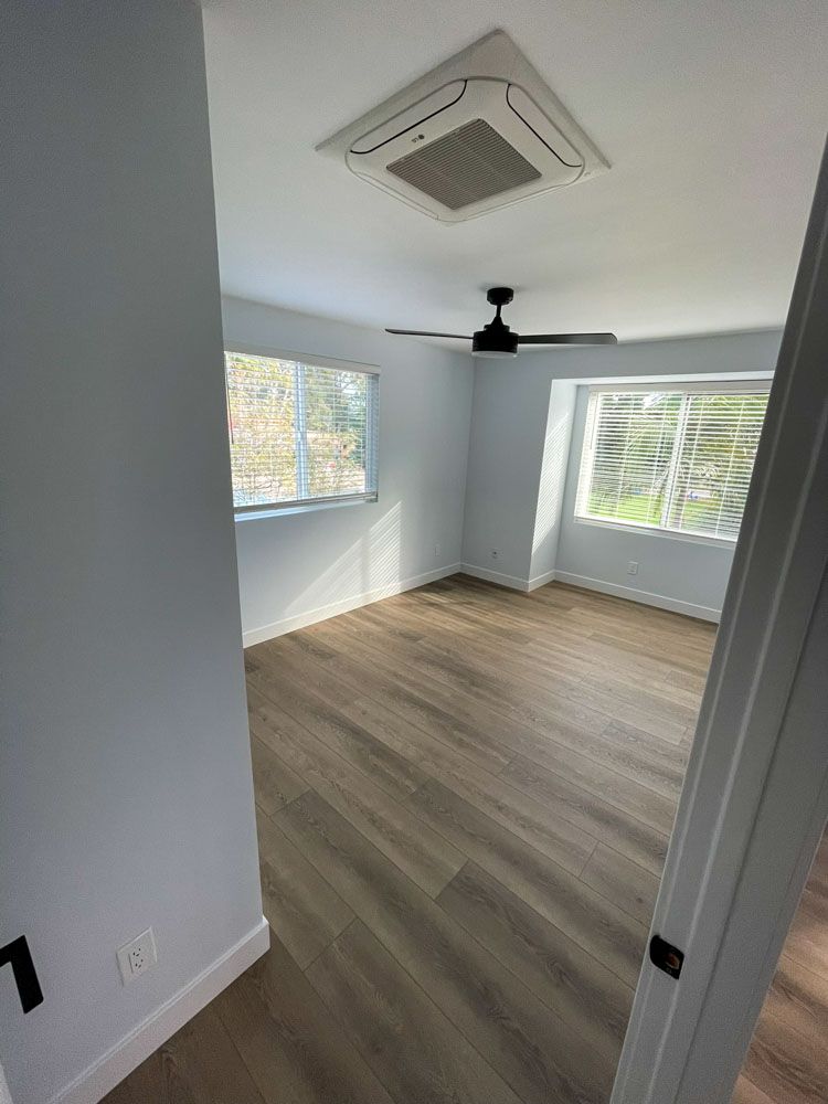 Empty bedroom with light blue walls, wooden floor, two windows, and a ceiling fan.