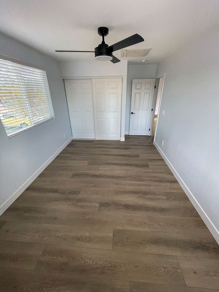 Empty bedroom with light wood-look flooring, white walls, and a black ceiling fan.  A closet and doorway are visible at the back.