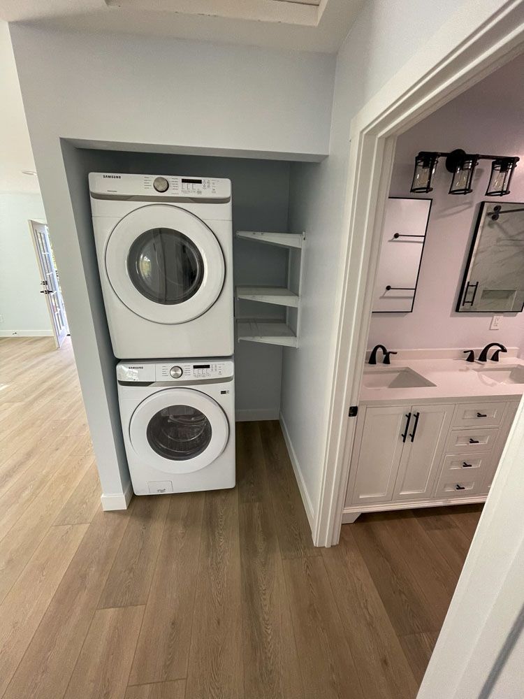 A stacked washer and dryer in a closet next to a bathroom. Light blue walls, wood floors, and white bathroom vanity.
