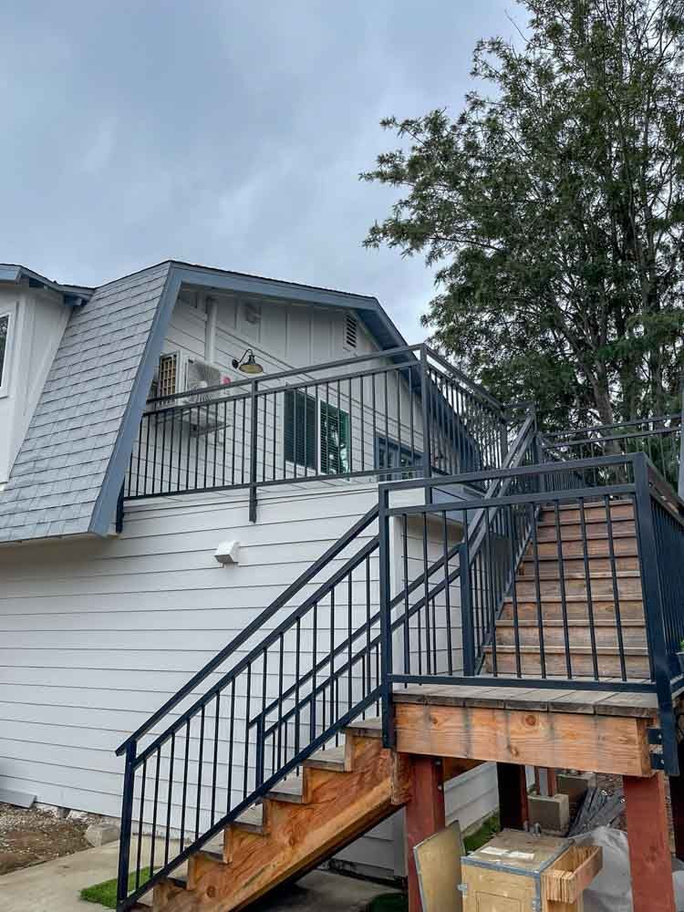 A two-story house with an outdoor staircase and a balcony. The house has gray roofing and white siding. The staircase has black metal railings.