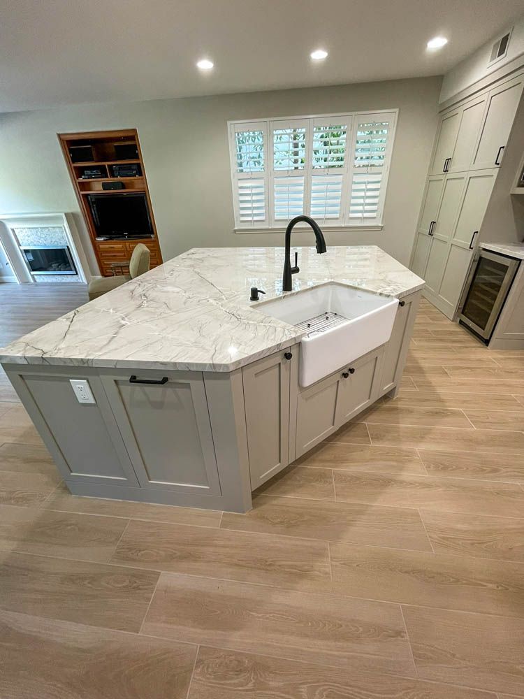 A newly renovated kitchen with a light gray island and white countertops. Features include a farmhouse sink, black faucet, and wooden floors.
