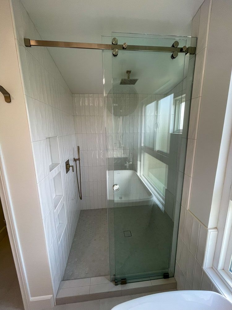 A modern bathroom with a glass shower enclosure, white tiled walls, and a sliding glass door. The shower has a built-in shelf and a bathtub.