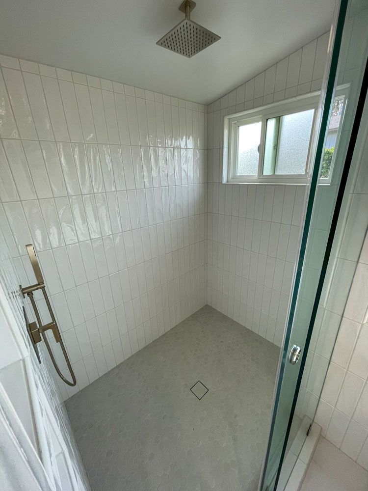 A modern, bright bathroom with white vertical tiles, grey floor, glass shower door, and gold shower fixtures. A small window is on the back wall.