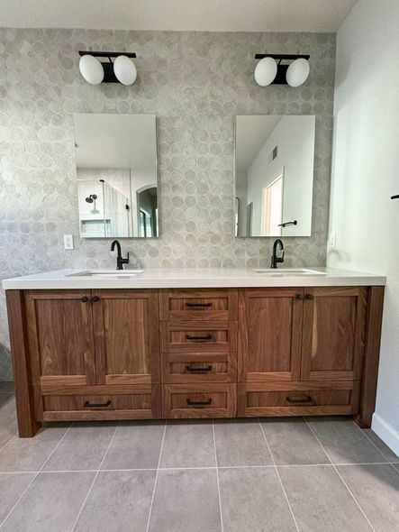 Bathroom with a wooden double vanity, gray countertops, and two mirrors. The backsplash is patterned gray tile, and the walls are white.
