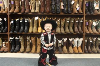 Young child in cowboy attire, posing in front of a wall of cowboy boots.