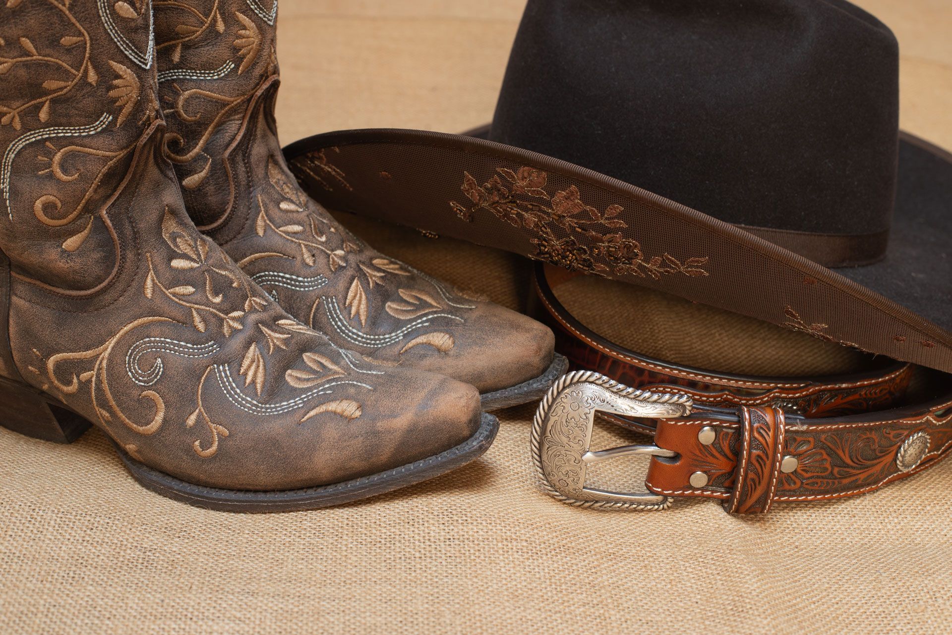 Cowboy boots, hat, and belt on a burlap surface. Brown, detailed leather with silver buckle.
