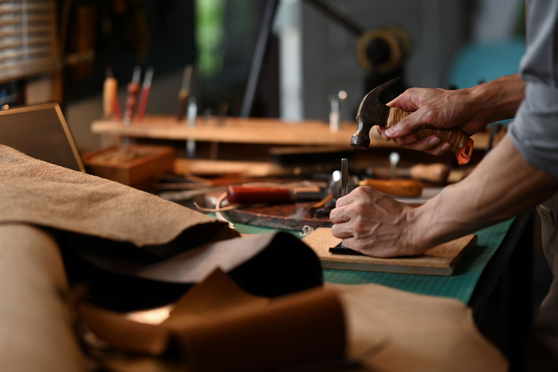 Hands hammering a small object on a wooden board; leather and tools on a workbench.