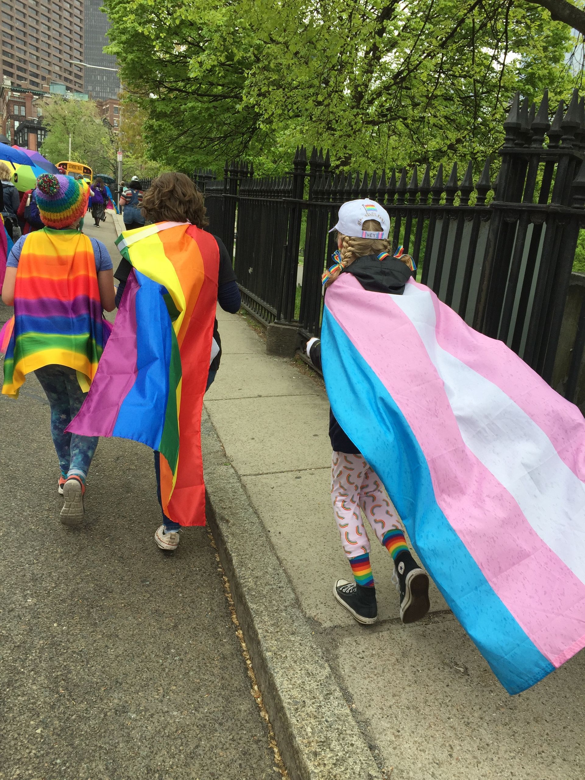 People walking on a sidewalk wearing rainbow and transgender pride flags as capes.