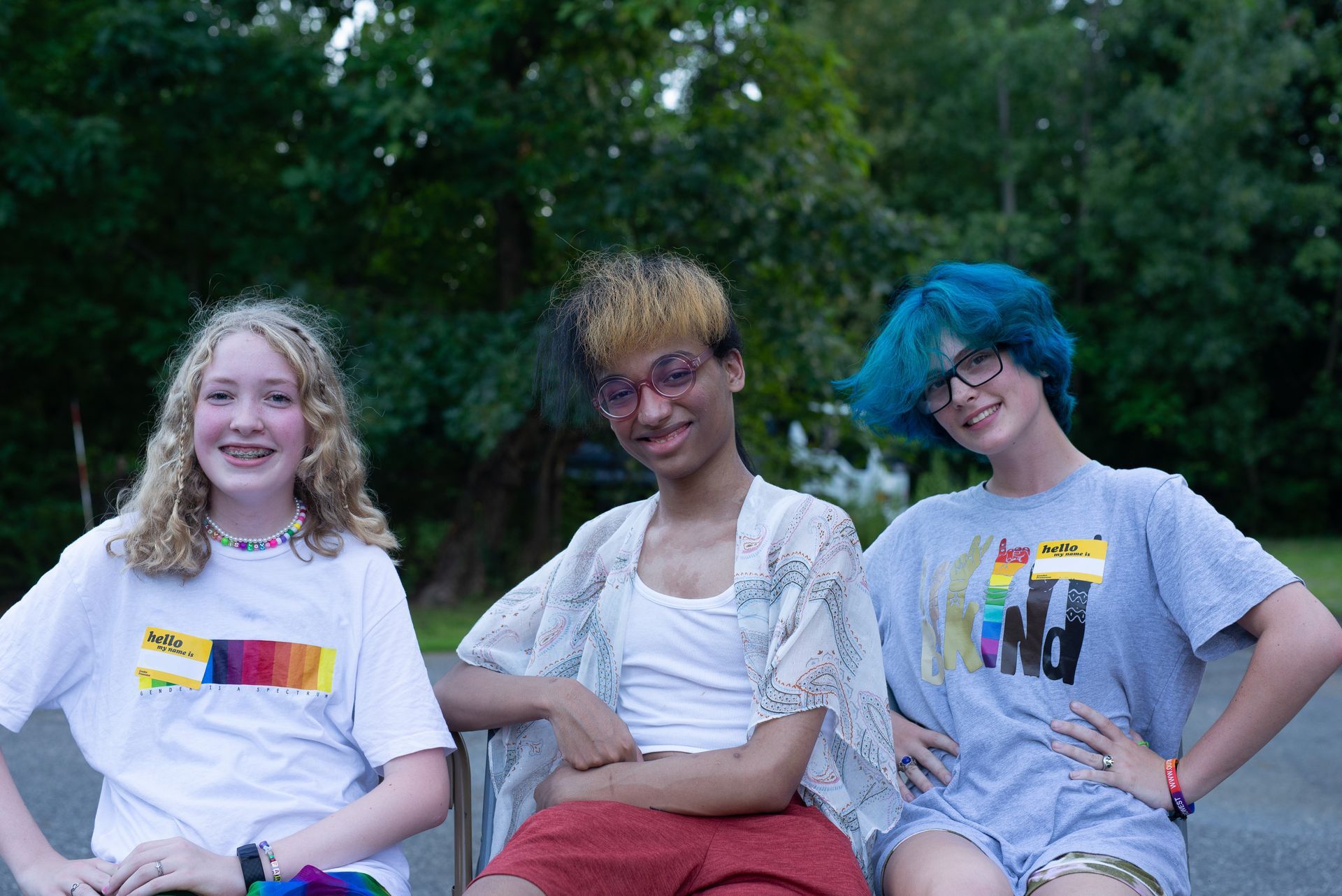 Three people smiling, sitting outside. Each wears a shirt with a rainbow design, and they have uniquely colored hair.