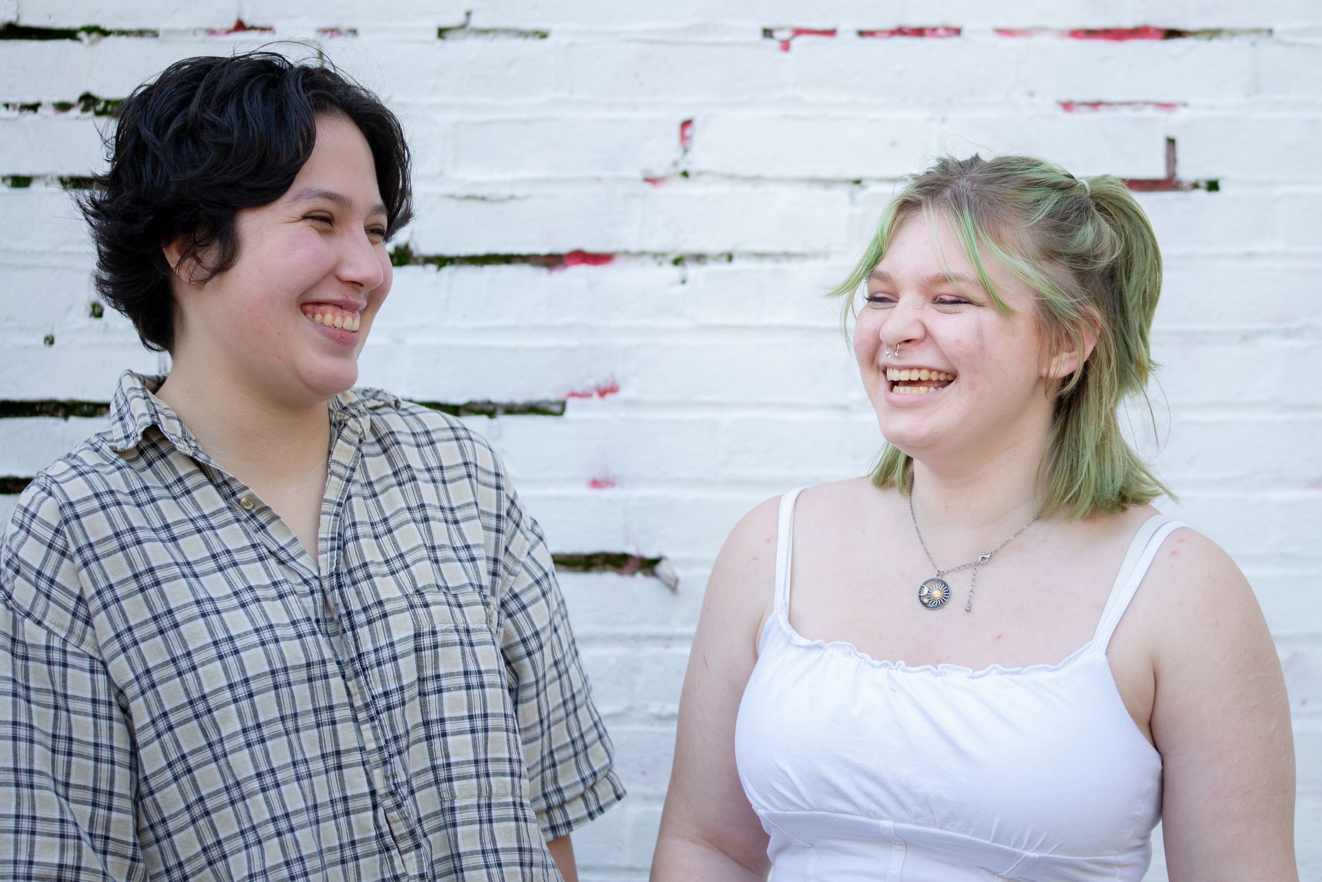 Two smiling people in front of a white brick wall. One wears a plaid shirt, the other a white tank top.