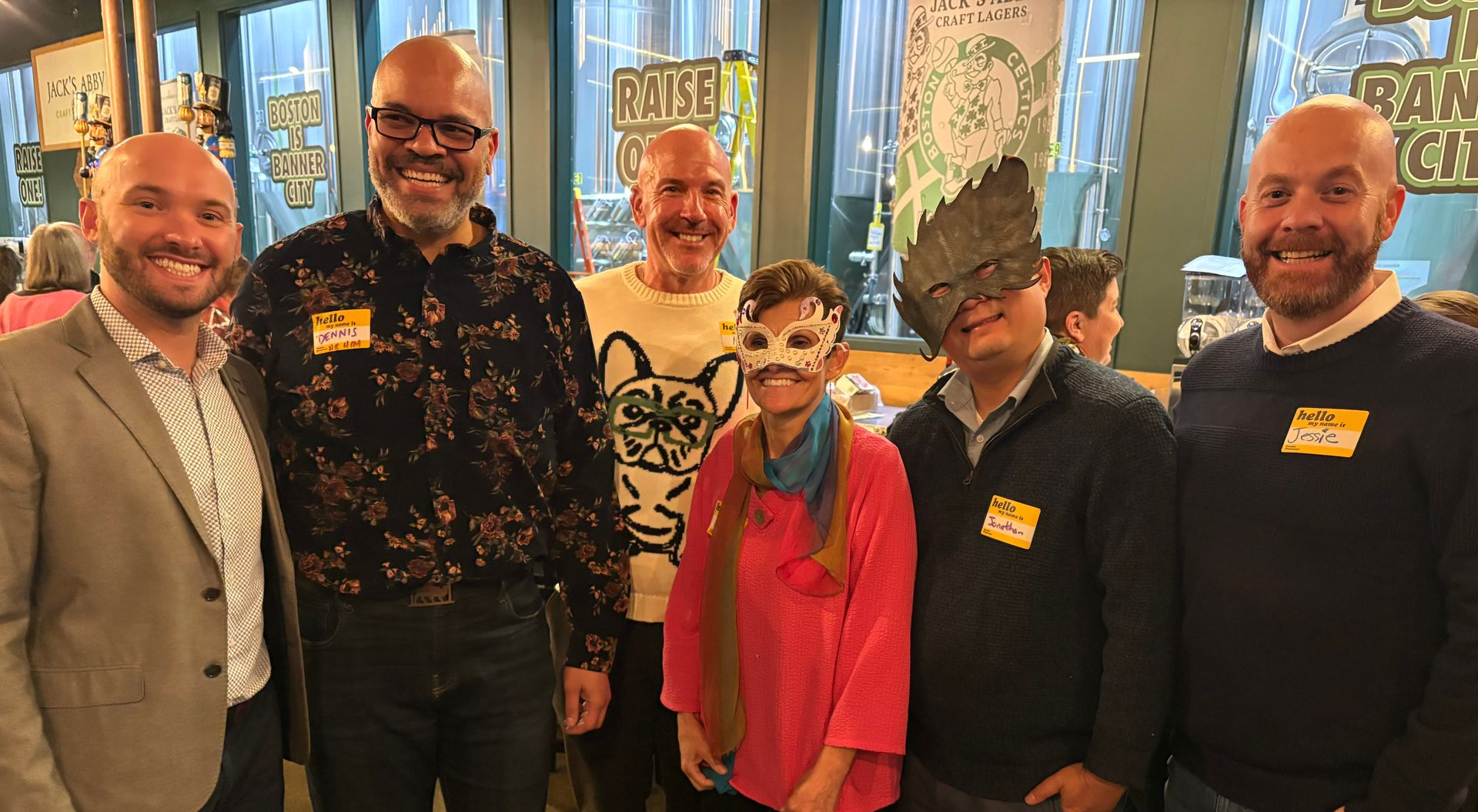 Group of six people smiling at a gathering, one wearing a mask, inside a building with a brewery backdrop.
