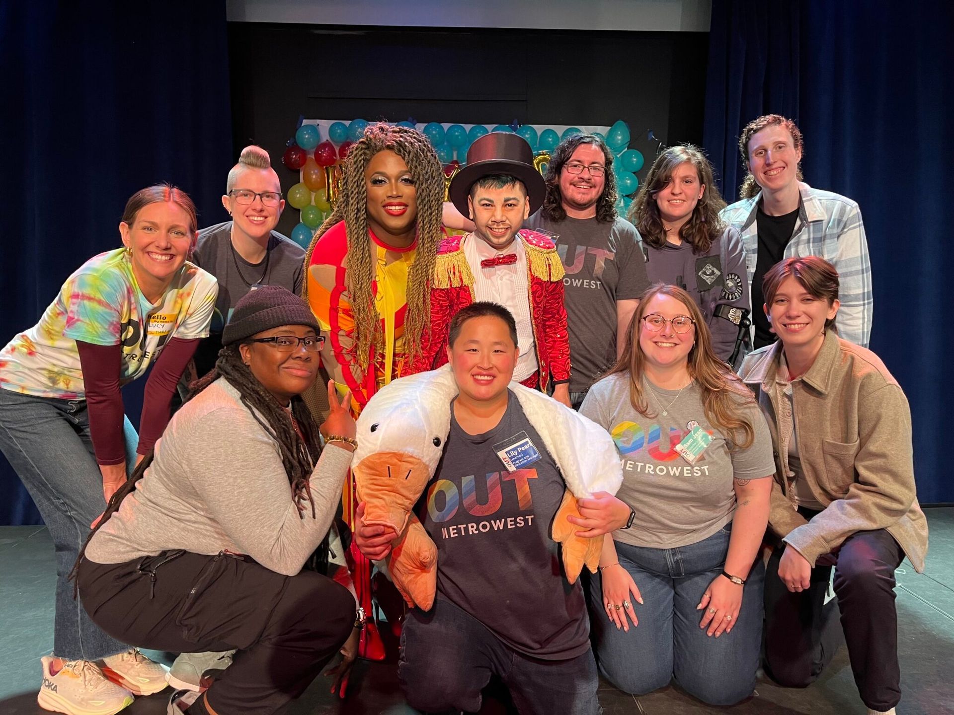 Group of people posing on a stage with balloons. Some are wearing colorful outfits and smiling.