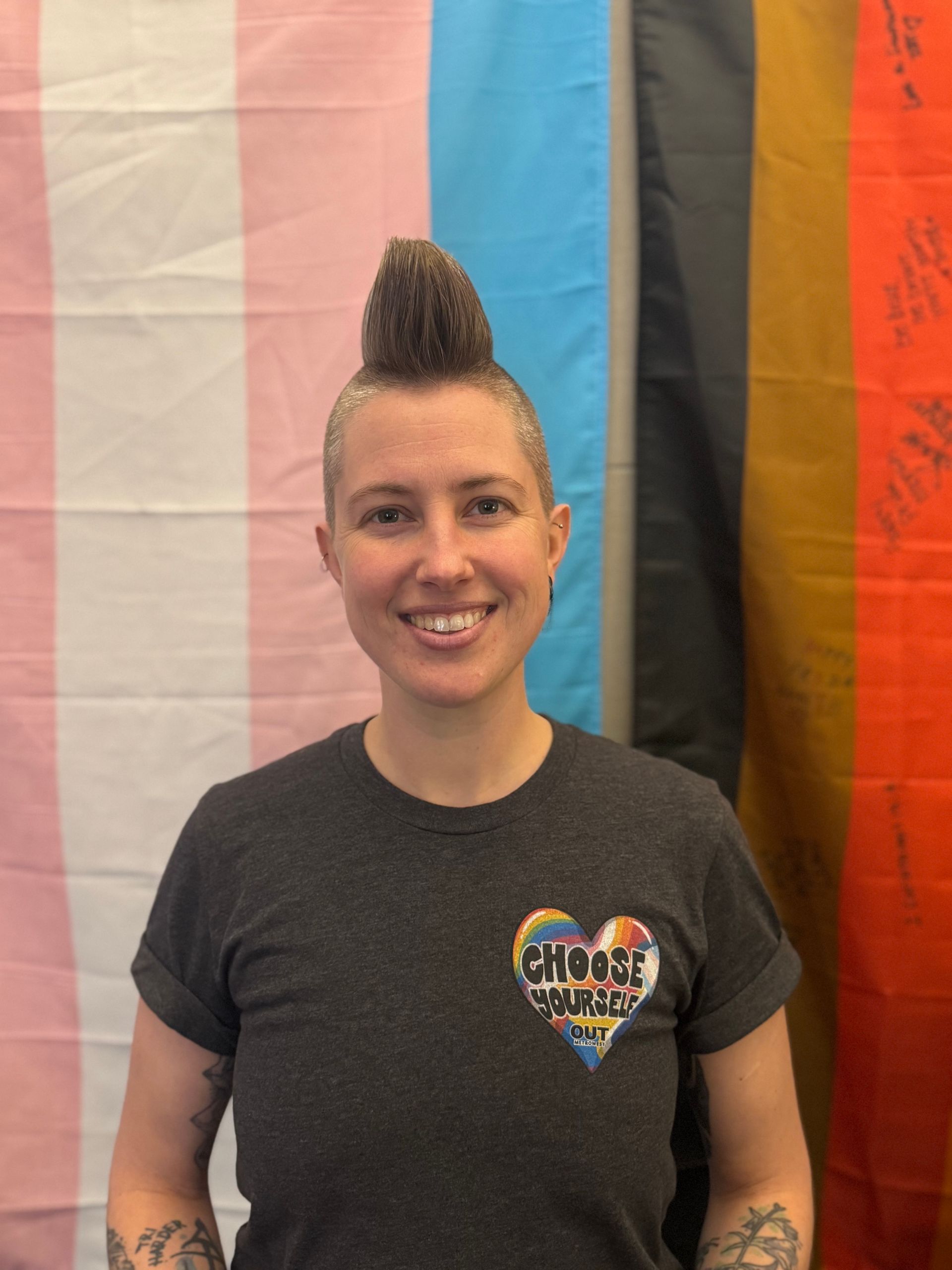 Person with a mohawk, smiling, wearing a t-shirt with a rainbow heart, in front of LGBTQ+ flags.