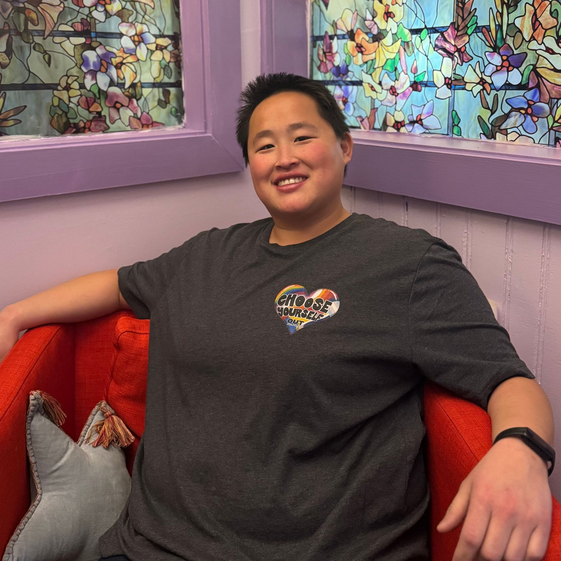 Person sitting on red couch, smiling, wearing a gray t-shirt with a heart-shaped logo. Purple walls, stained glass window.