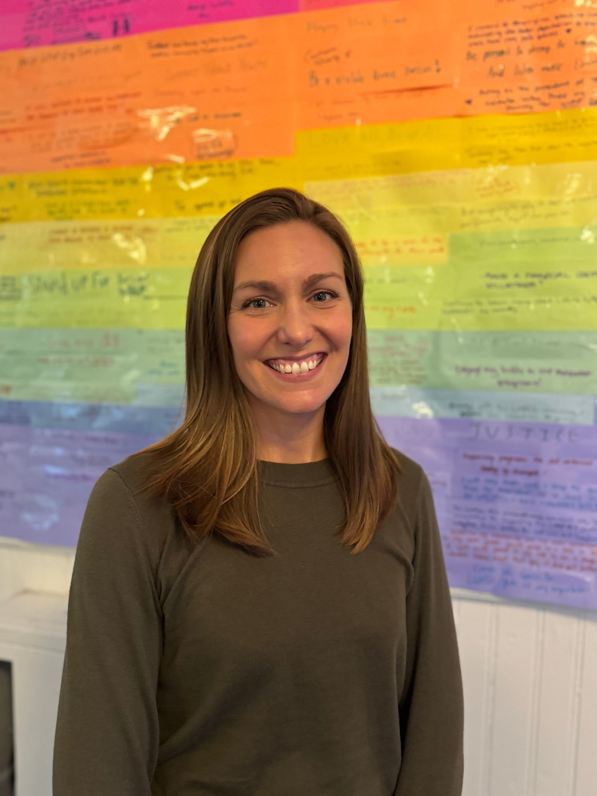 Person smiling in front of a colorful, handwritten backdrop. She has long brown hair and is wearing a green shirt.