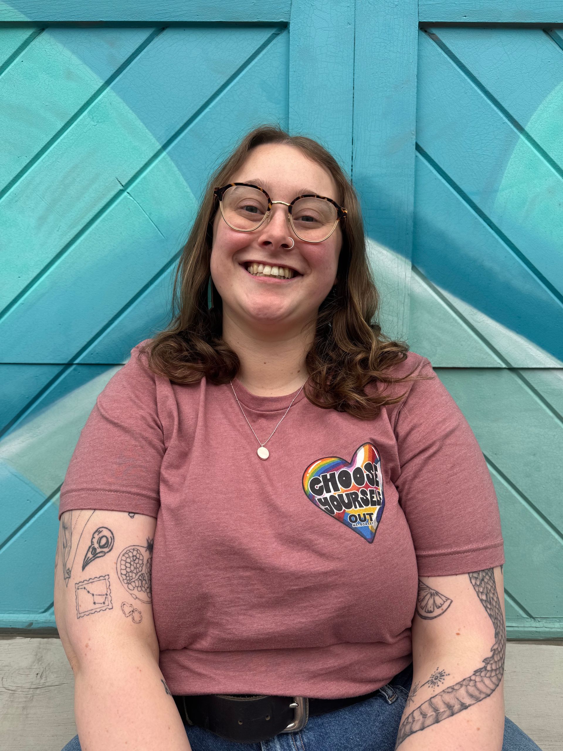 Person smiling in front of a turquoise door, wearing glasses, and a pink shirt with a heart patch and arm tattoos.