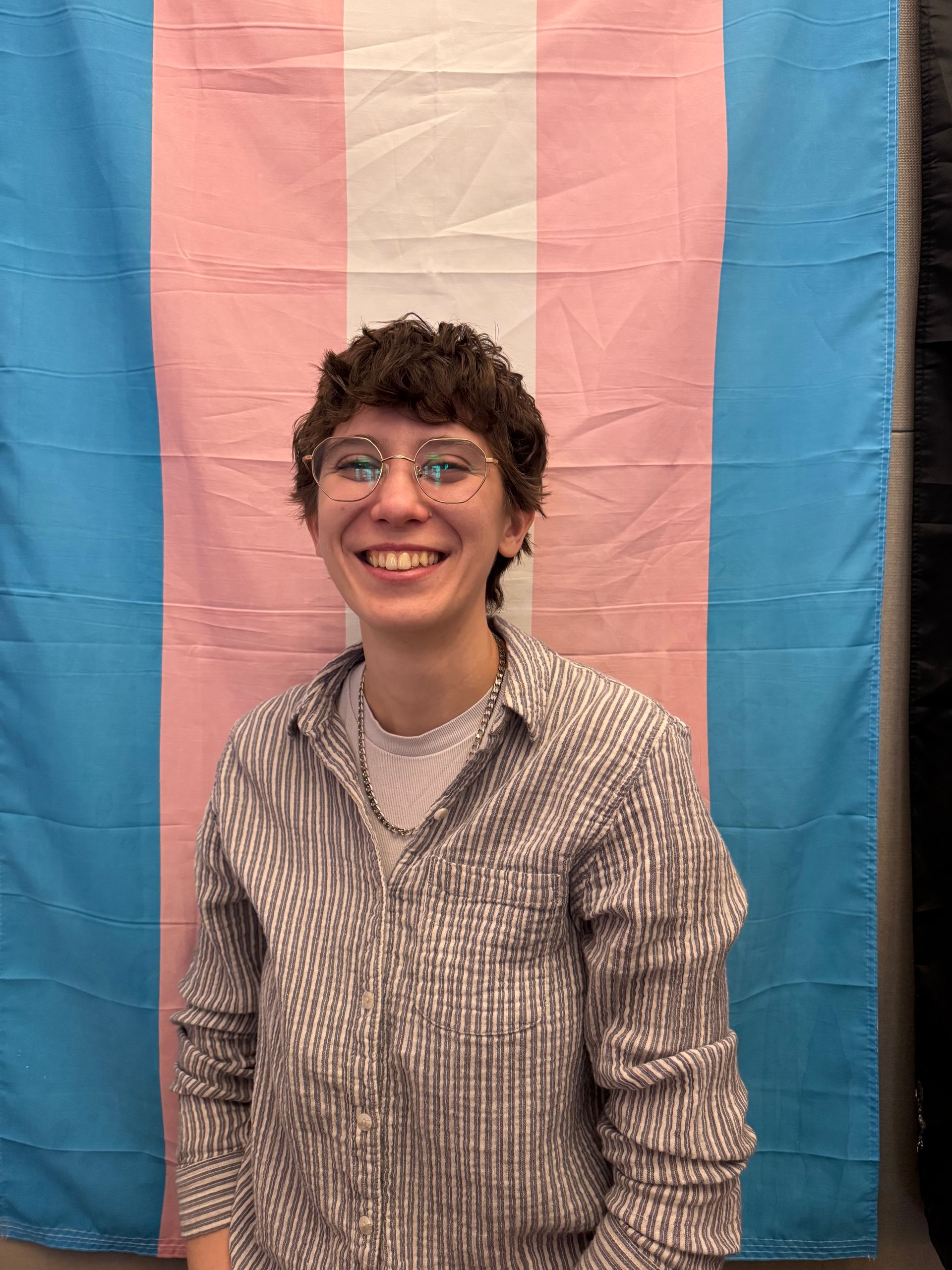 Person smiling in front of a transgender flag. They wear glasses and a striped shirt.