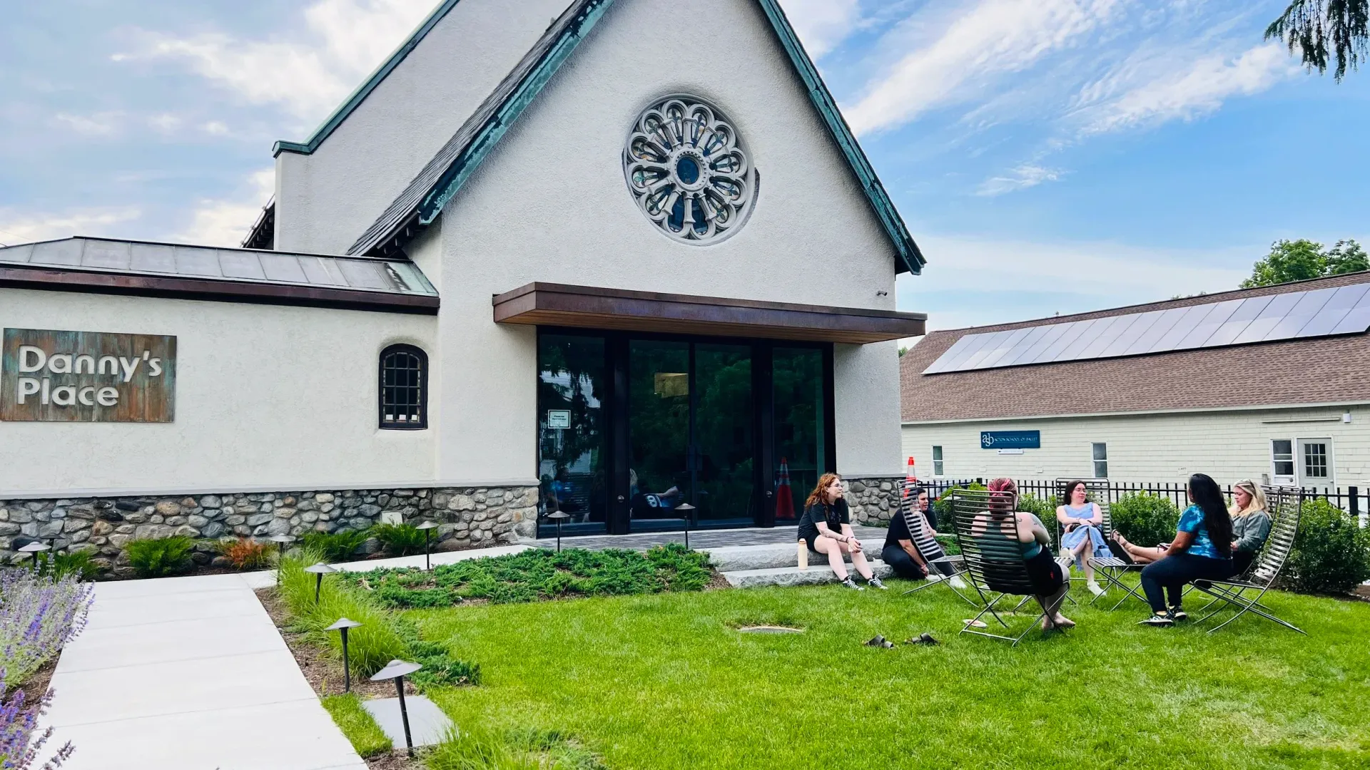 Exterior of Dannys's Place, people seated on chairs in front of building with rose window and solar panels.