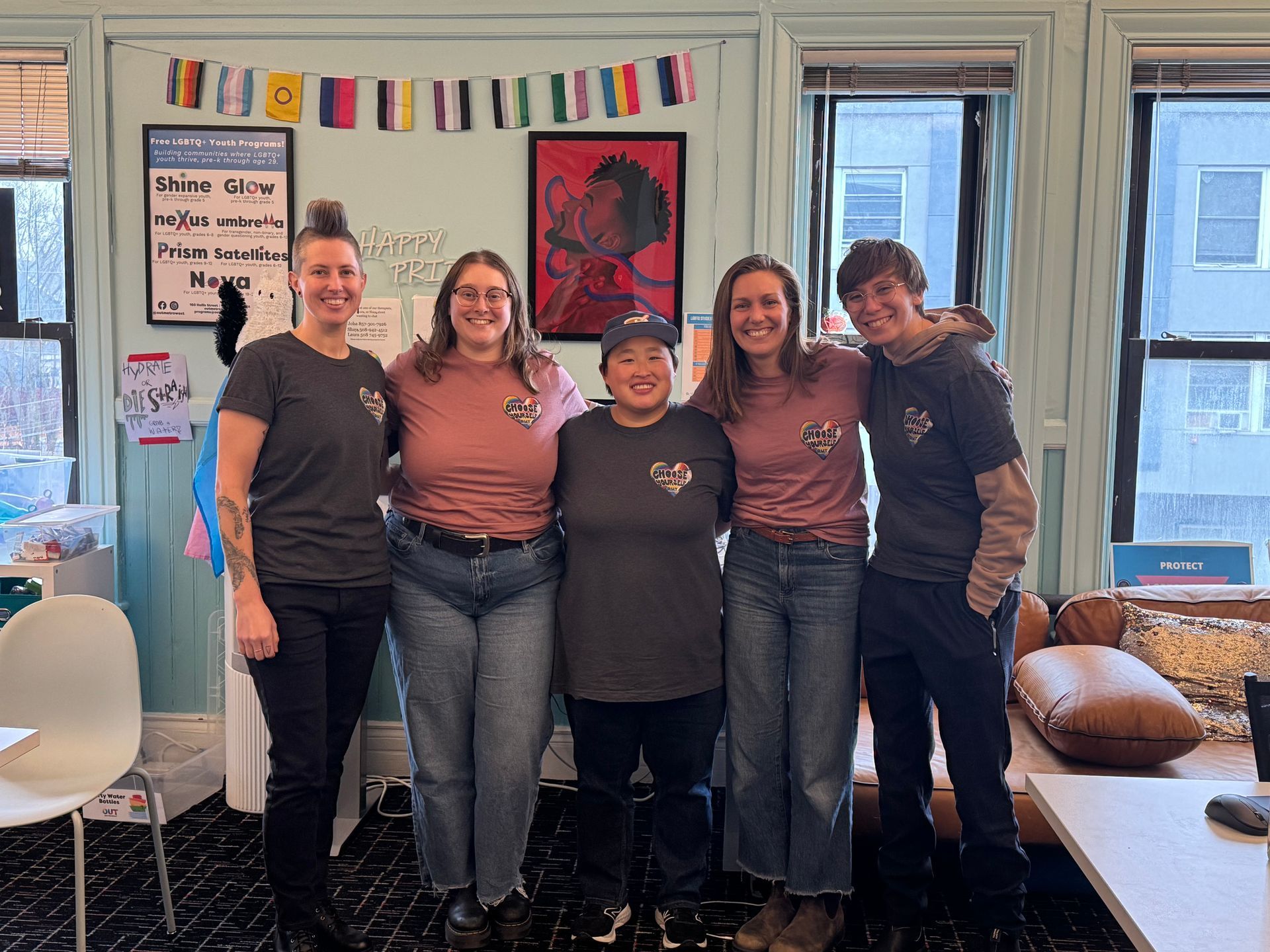 Five people smiling, arms around each other. Inside, with flags and a poster on the wall. They wear t-shirts.