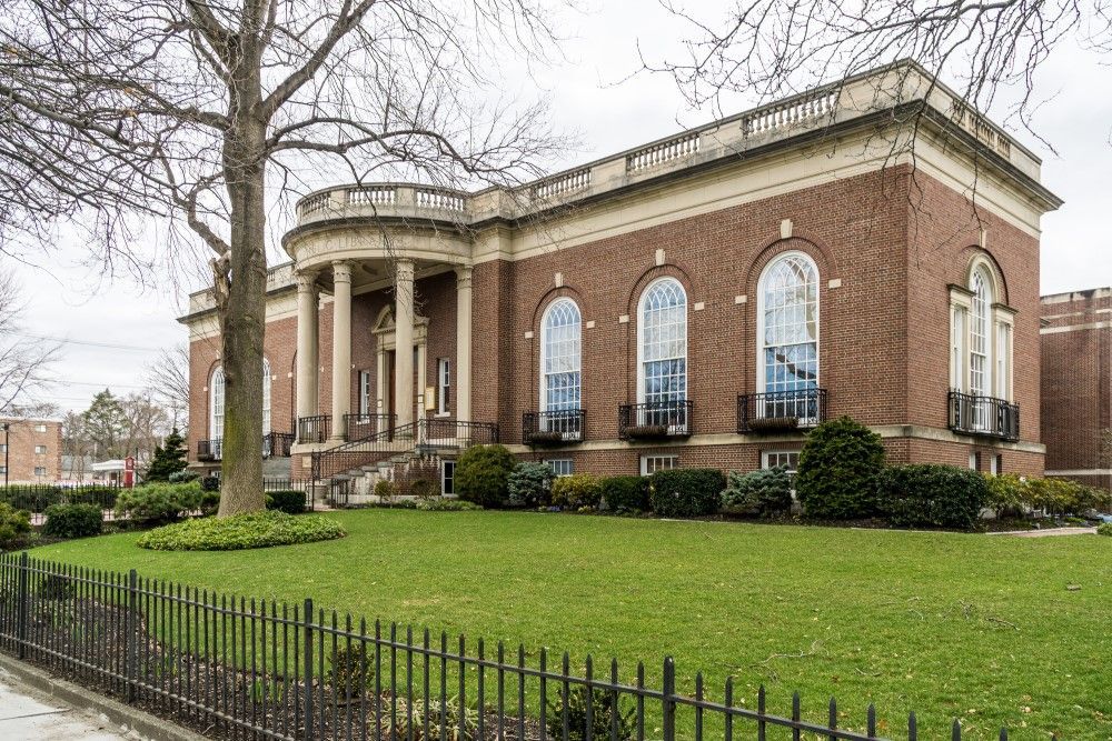Exterior of the Waltham Public Library, a brick building with arched windows, and a green lawn behind a wrought iron fence.