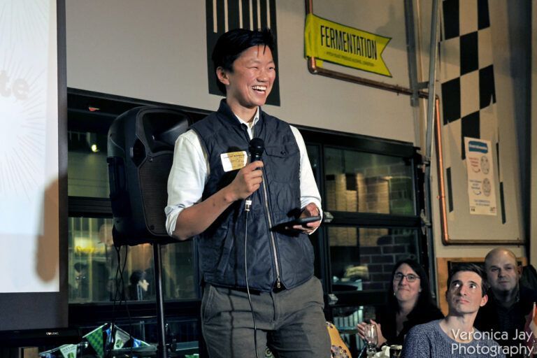 Person speaking at event, holding microphone and phone, smiles.