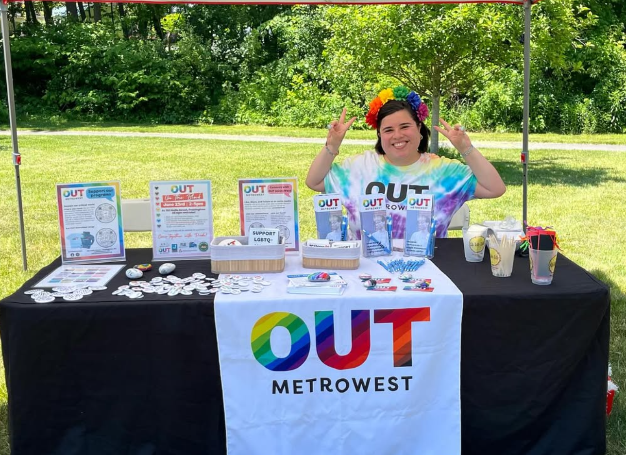 Person at OUT MetroWest table, wearing rainbow hat, making peace signs, in park.
