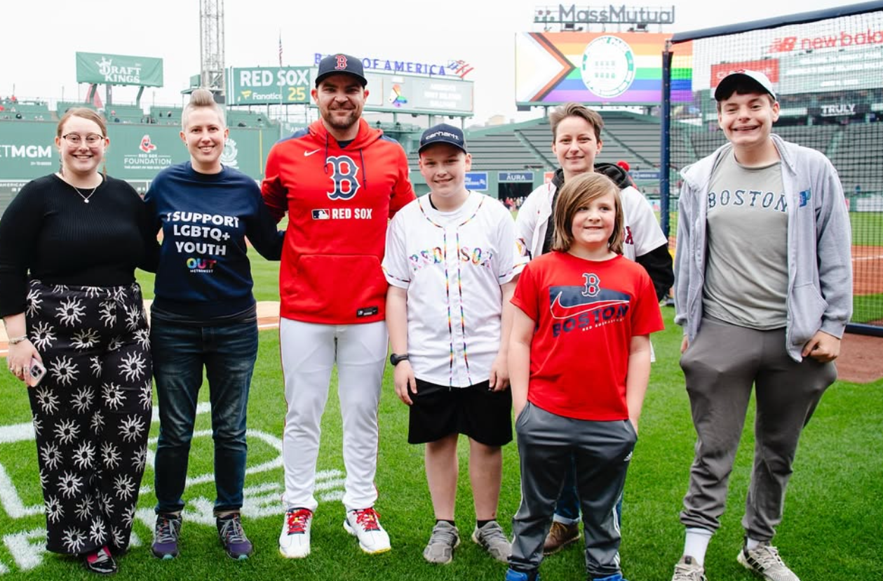 Group of people smiling on baseball field. Red Sox player stands with fans. Pride flag visible in background.